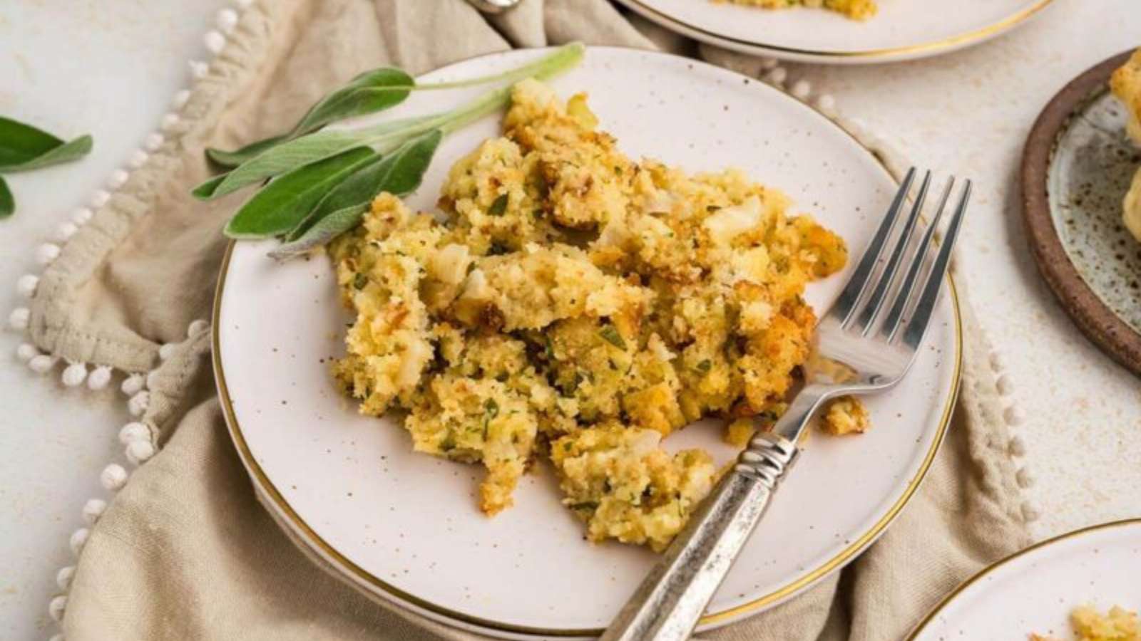 A plate of stuffing garnished with fresh sage, accompanied by a fork, resting on a beige napkin.