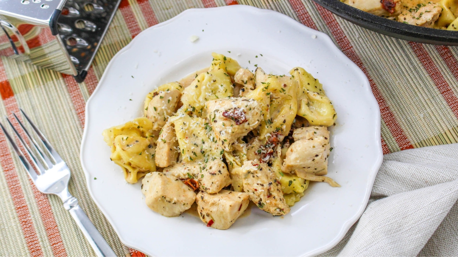 A white plate with creamy tortellini and chunks of seasoned chicken, garnished with herbs. A fork, napkin, and cheese grater are nearby on a striped cloth.