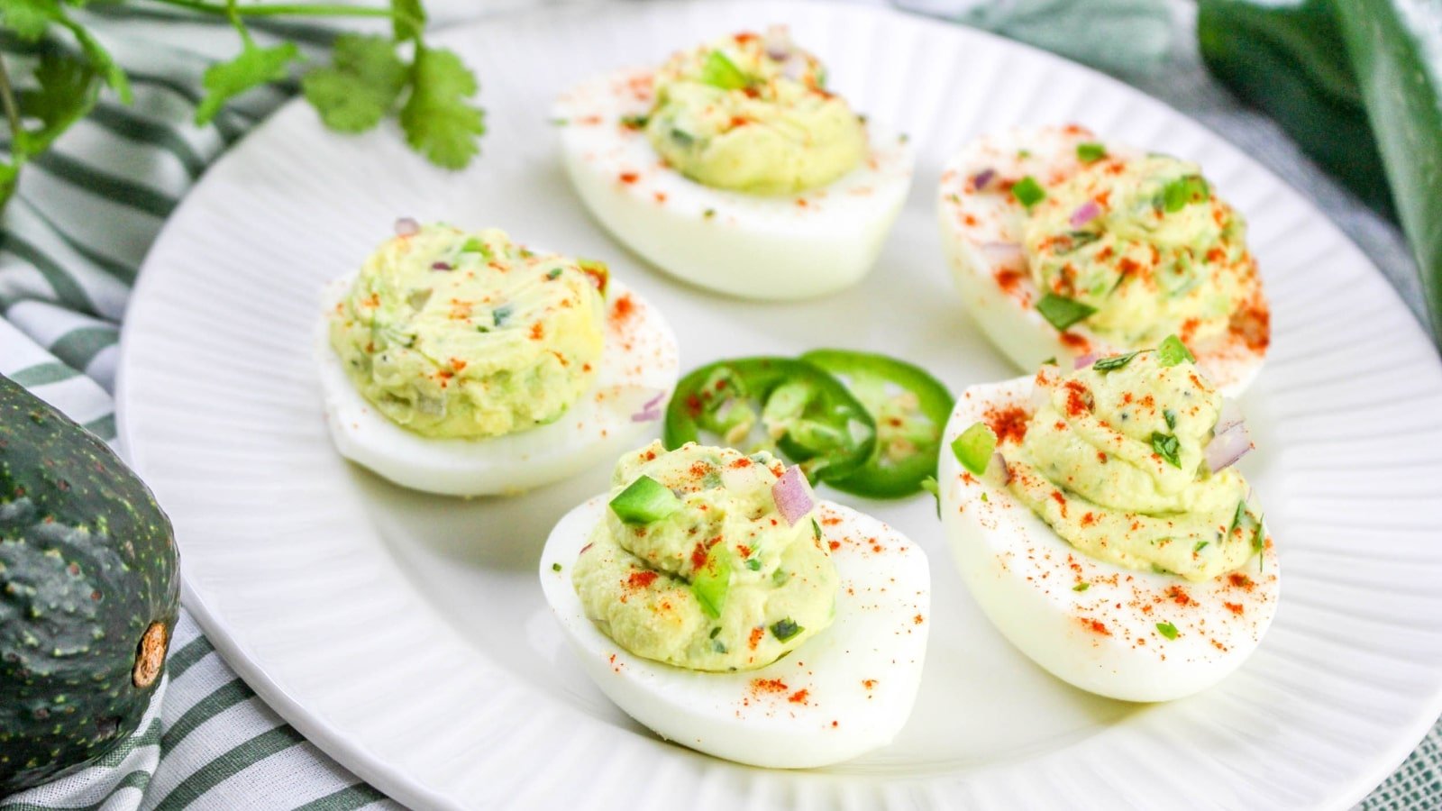 Plate of deviled eggs with avocado filling, garnished with paprika and green herbs, and sliced jalapeño.