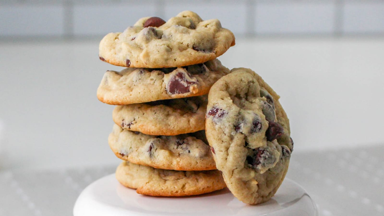 A stack of five chocolate chip cookies with one leaning against them on a white surface.