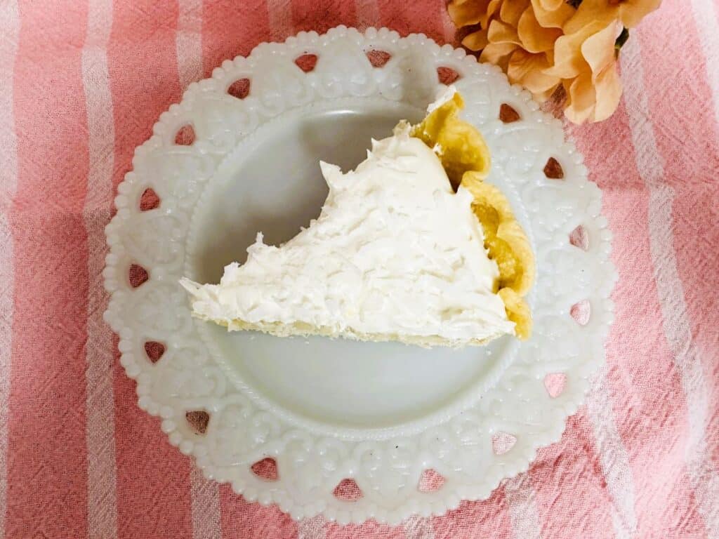A slice of cream pie with a golden crust on a decorative white plate, placed on a pink striped tablecloth. A peach-colored flower is in the corner.
