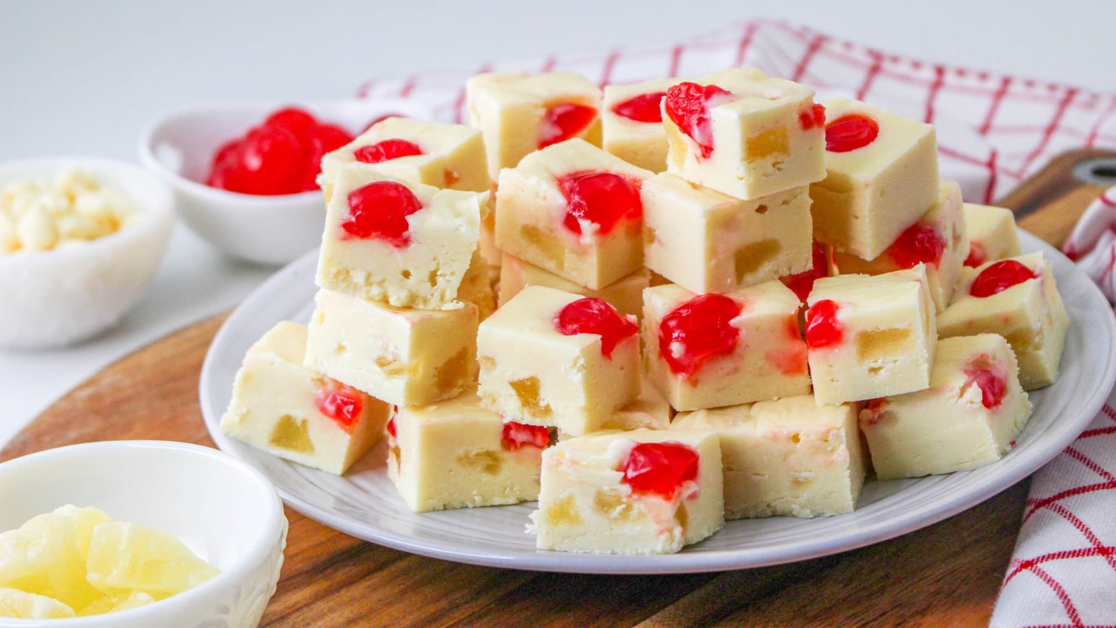 A plate of white fudge pieces topped with red cherries, surrounded by small bowls of ingredients on a wooden surface with a red checkered cloth in the background.