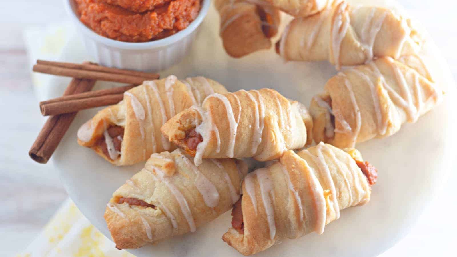 A plate of drizzled croissant rolls with a side of cinnamon sticks and a dish of spreadable orange paste.