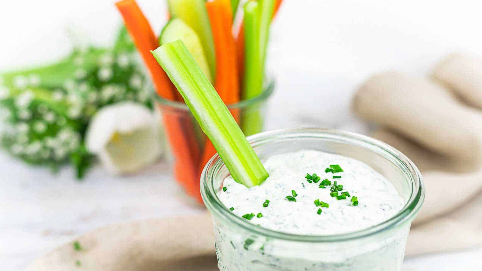 Glass jar with creamy dip garnished with chives, a celery stick in it. Blurred background shows a jar with carrot, celery, and cucumber sticks, and a white flower.