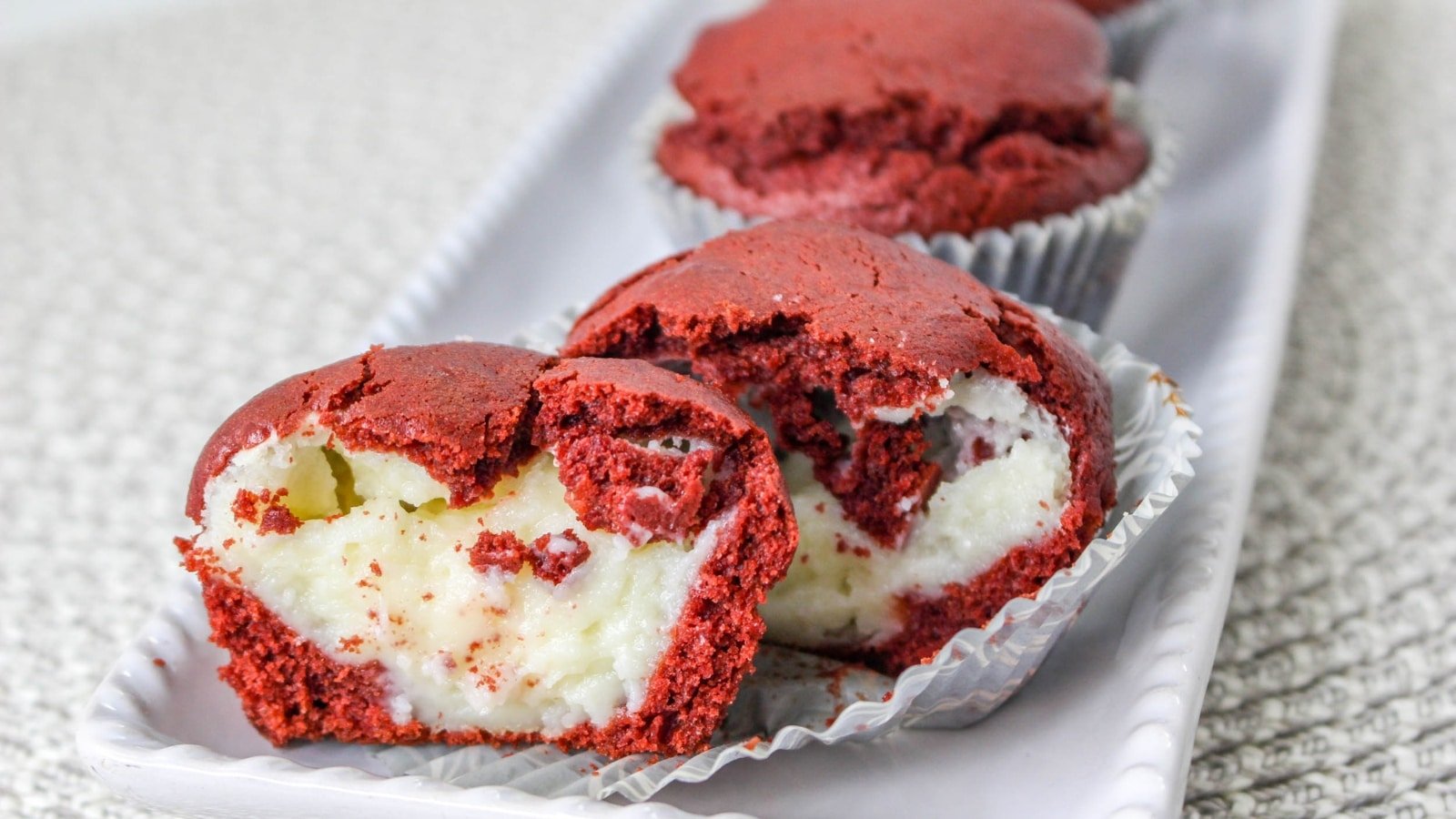 Red velvet cupcakes filled with cream cheese frosting, one cut open to reveal the filling, on a white platter.