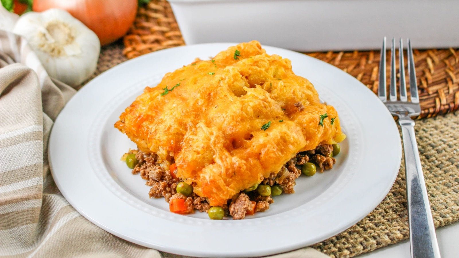 A plate of shepherds pie topped with melted cheese, showing layers of ground meat, peas, and carrots. Near the plate are a fork, garlic, onion, and a striped napkin.