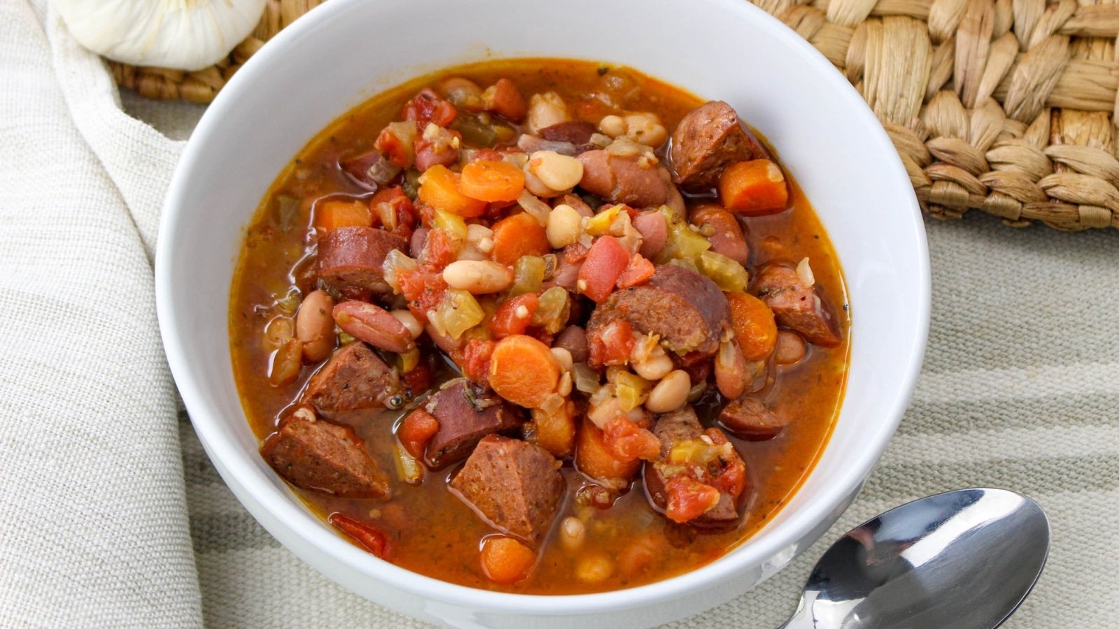 A bowl of hearty bean and sausage stew with carrots and tomatoes, placed on a woven mat next to a spoon and a cloth napkin.