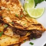 A close-up of a quesadilla filled with shredded meat and garnished with chopped herbs, served with lime wedges on the side.