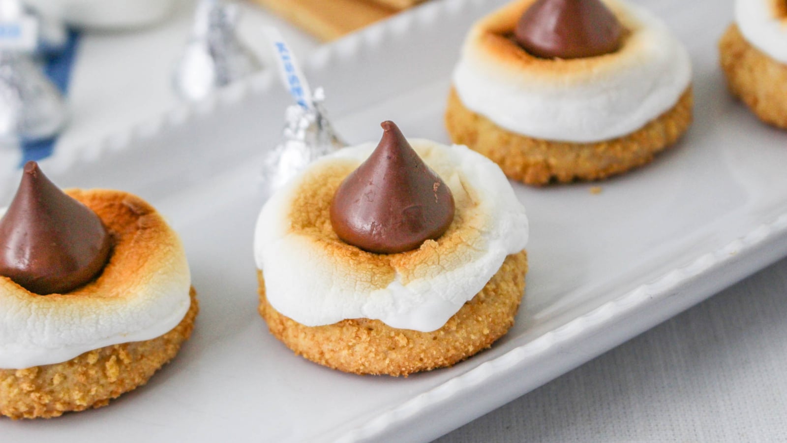 Three smores cookies with toasted marshmallow and chocolate on a tray, with foil-wrapped chocolates in the background.