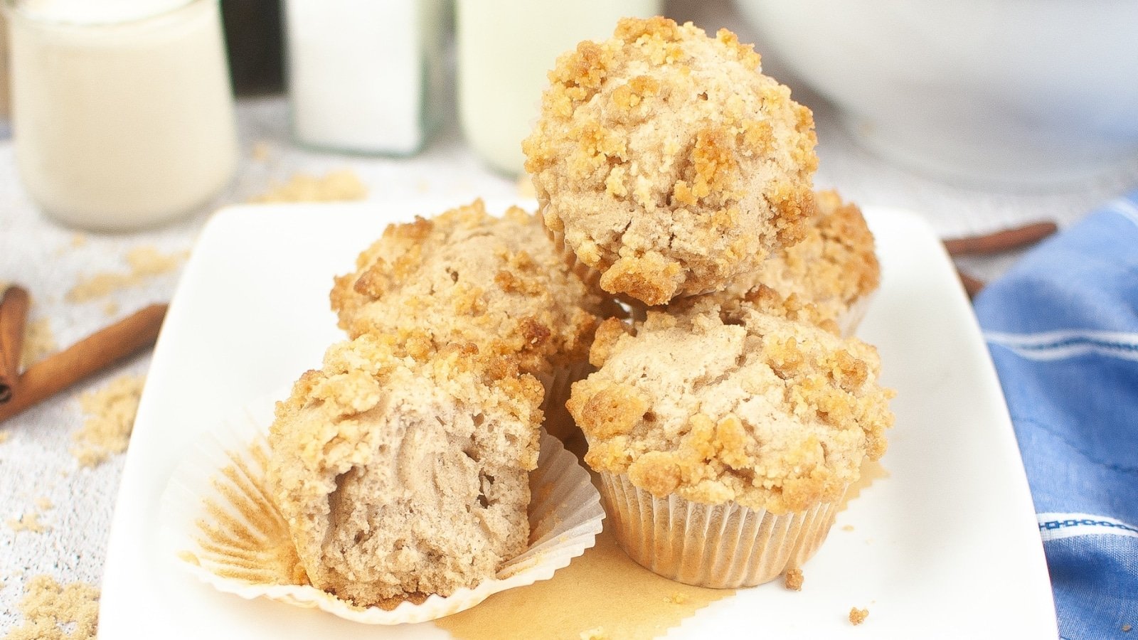 A plate of five cinnamon muffins with crumbly tops; one muffin partially unwrapped and cut in half.