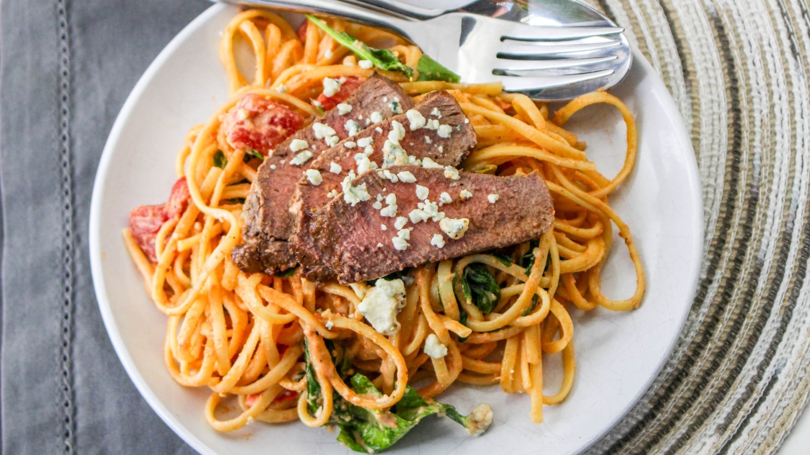 Plate of spaghetti with sliced steak, spinach, tomatoes, and crumbled cheese, served with a fork and spoon on a round, white plate.