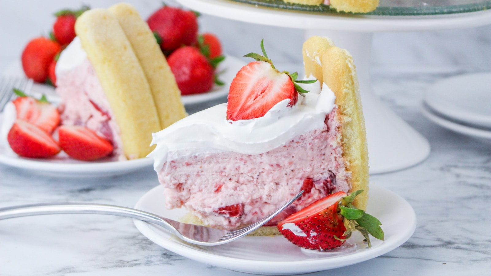 A slice of strawberry shortcake topped with whipped cream and a strawberry slice, surrounded by ladyfingers. Fresh strawberries and a fork are on the plate.