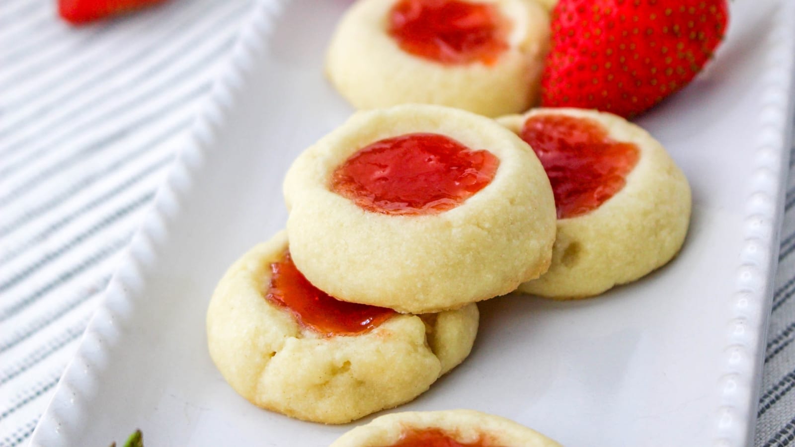 A white plate with strawberry jam thumbprint cookies, placed on a striped napkin.