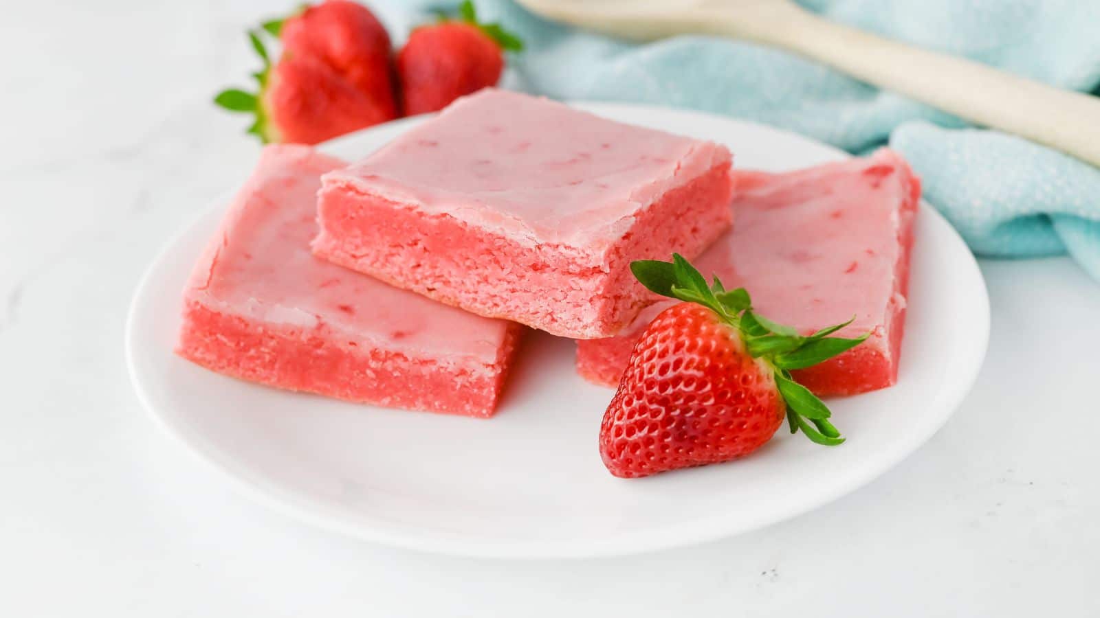 Three slices of pink strawberry dessert on a white plate, garnished with fresh strawberries. A wooden spoon and a blue cloth are in the background.