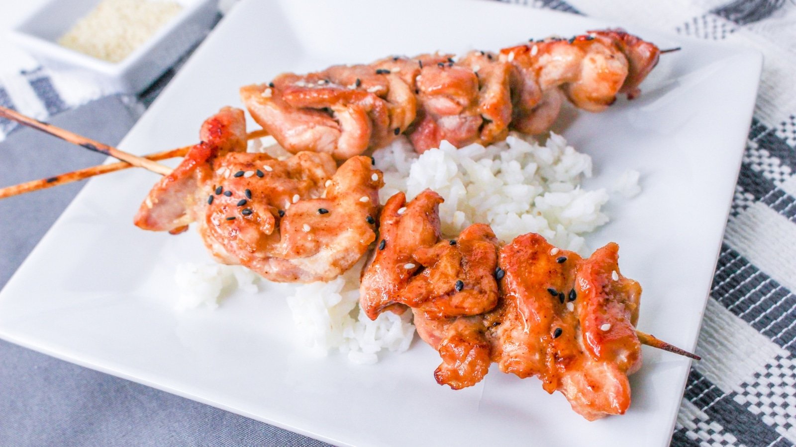 Two grilled chicken skewers with sesame seeds on white rice, served on a square white plate.