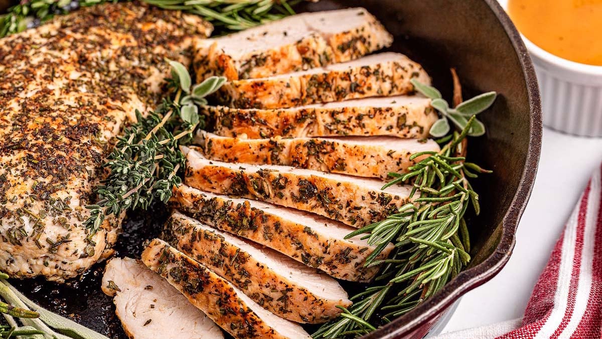 Sliced herb-crusted turkey tenderloin in a skillet, garnished with sprigs of rosemary and sage, with a gravy dish nearby.