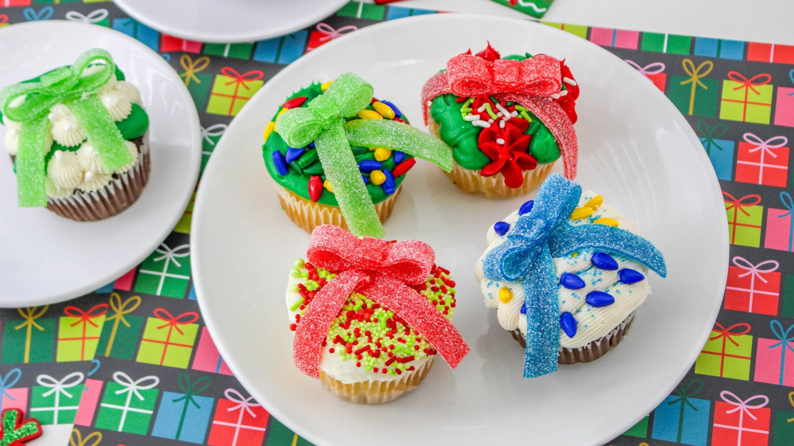 Four cupcakes decorated as colorful gift boxes with frosting and sugar bows, on a white plate. The background has a pattern of variously colored present illustrations.