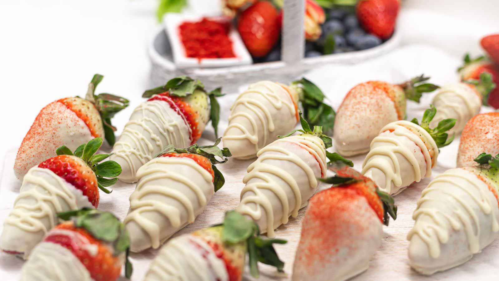 Fresh strawberries coated in white chocolate and drizzled with more chocolate are arranged on a white surface. Some are garnished with red sprinkles. A basket of mixed berries is in the background.