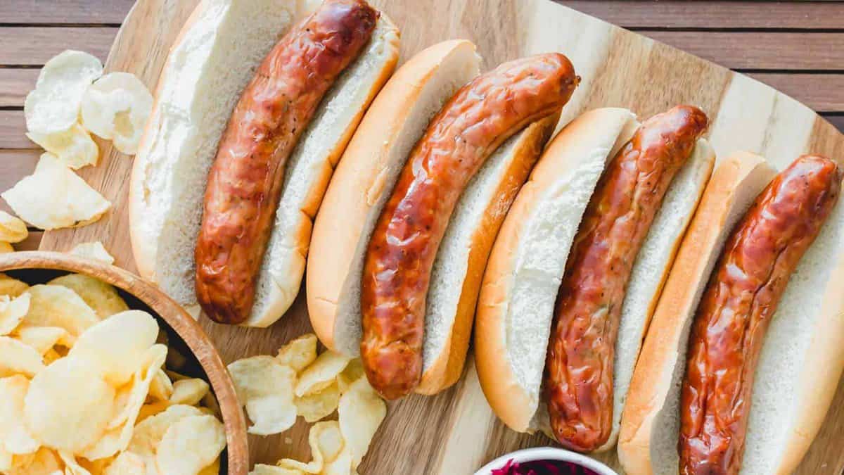 Four grilled sausages in buns are on a wooden board, surrounded by potato chips and a bowl of coleslaw.