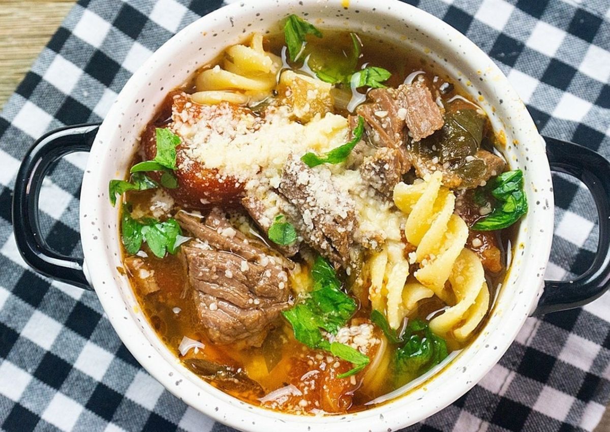 A bowl of beef and vegetable soup with pasta, topped with grated cheese and fresh parsley, sits on a black-and-white checkered cloth—one of our 14 Easy Beef Dishes you’ll want to make again and again.