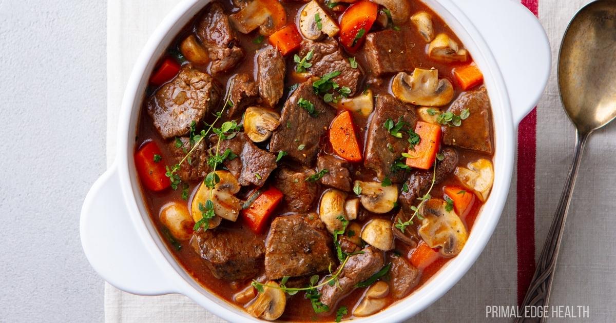 A white bowl filled with beef stew containing chunks of beef, mushrooms, carrots, and garnished with herbs. A spoon is placed on the right side on a cloth.