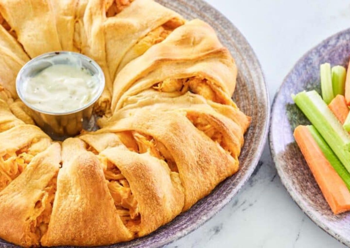 A plate with a ring-shaped baked pastry, inspired by 12 Delicious Dishes Using Crescent Rolls, filled with shredded chicken and a cup of dipping sauce in the center, served alongside carrot and celery sticks.