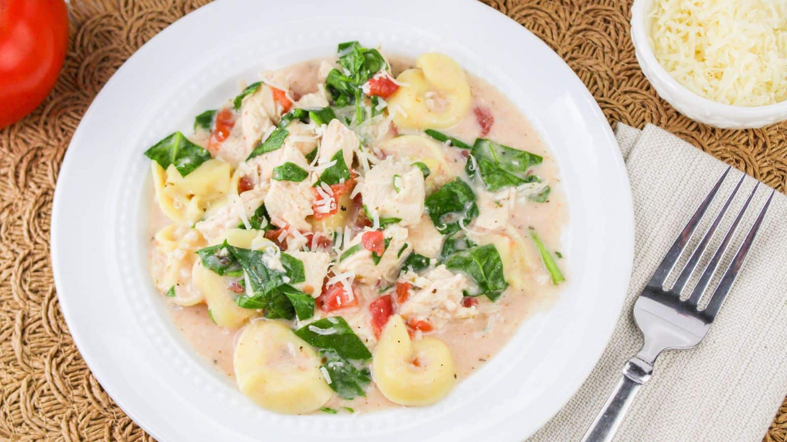 A white plate with creamy tortellini, shredded chicken, spinach, and diced tomatoes. A fork and a small bowl of grated cheese are on the side.
