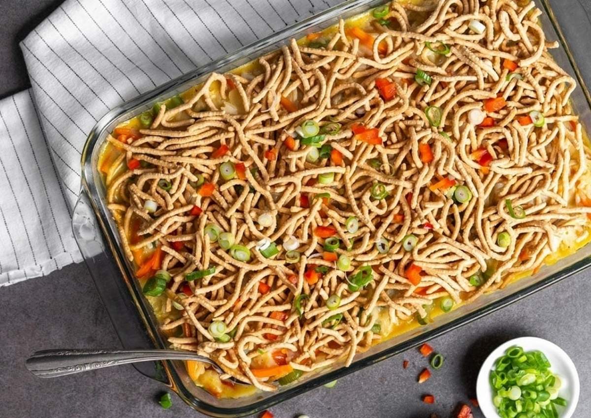 A rectangular glass baking dish filled with a noodle casserole, inspired by 12 Simple Casserole Recipes, is topped with crispy chow mein noodles, chopped green onions, and diced red bell peppers, with a metal spoon on the side.