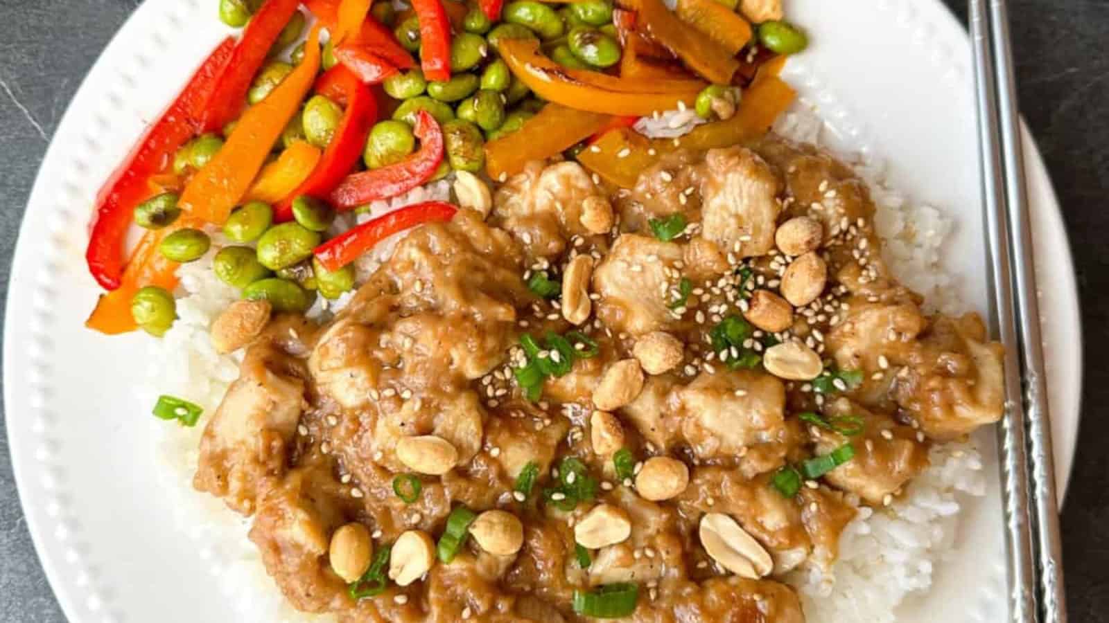 A white plate with rice topped with peanut chicken, garnished with chopped nuts and green onions, and a side of mixed vegetables including red peppers, carrots, and edamame. Chopsticks are on the side.