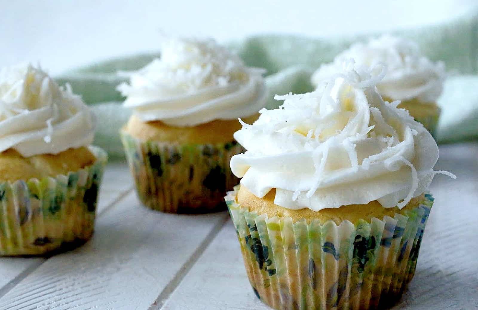 Four cupcakes with white frosting and coconut shavings are arranged on a white wooden surface.
