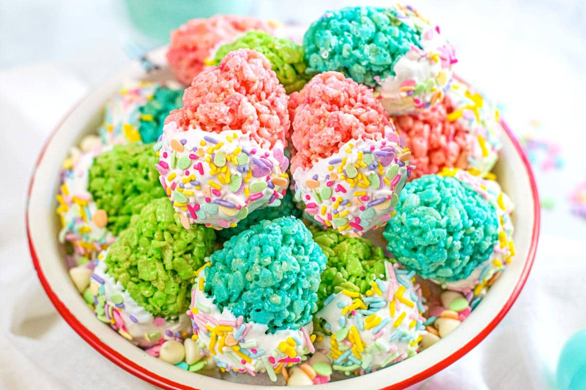 A bowl of colorful rice crispy treats, dipped in white icing and decorated with sprinkles, in shades of pink, blue, and green.