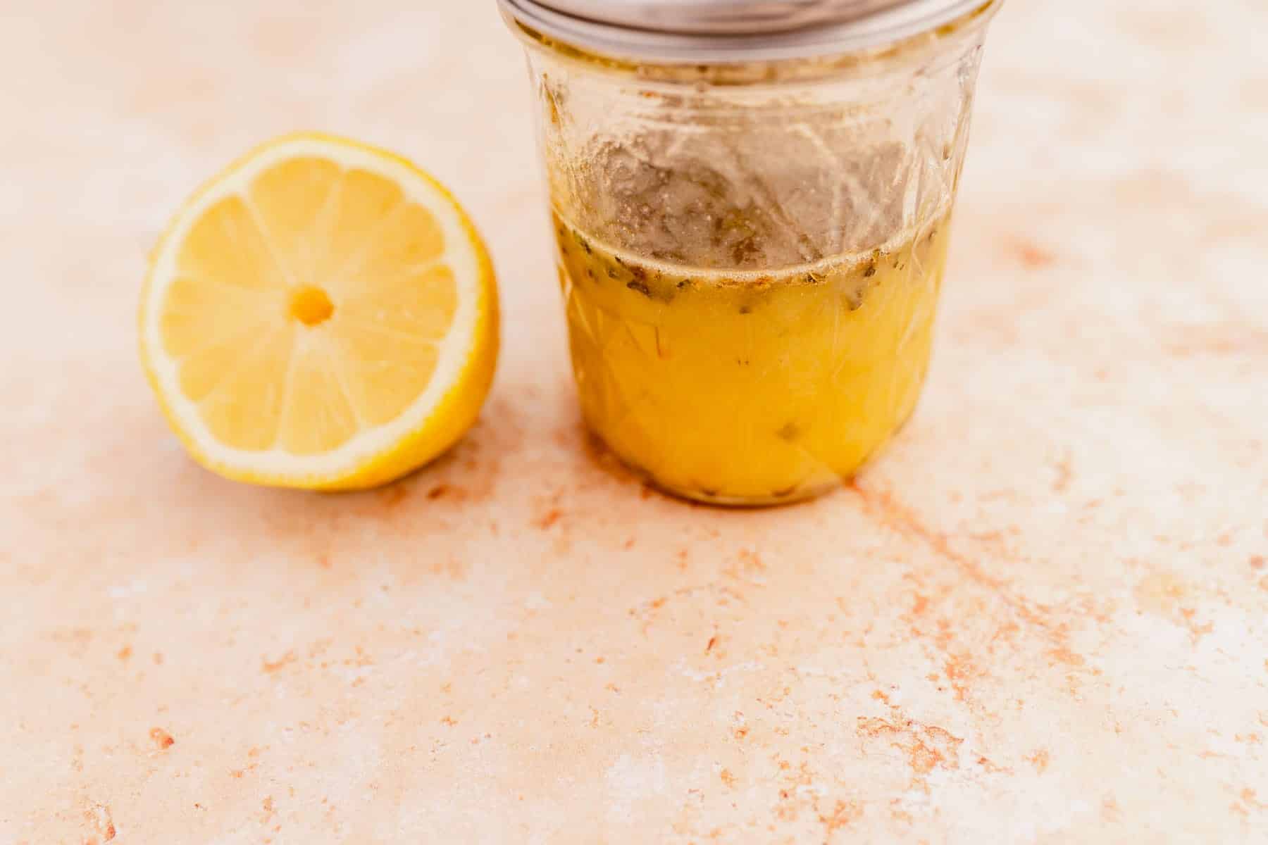 A half lemon and a glass jar with yellow liquid sit on a textured beige surface.