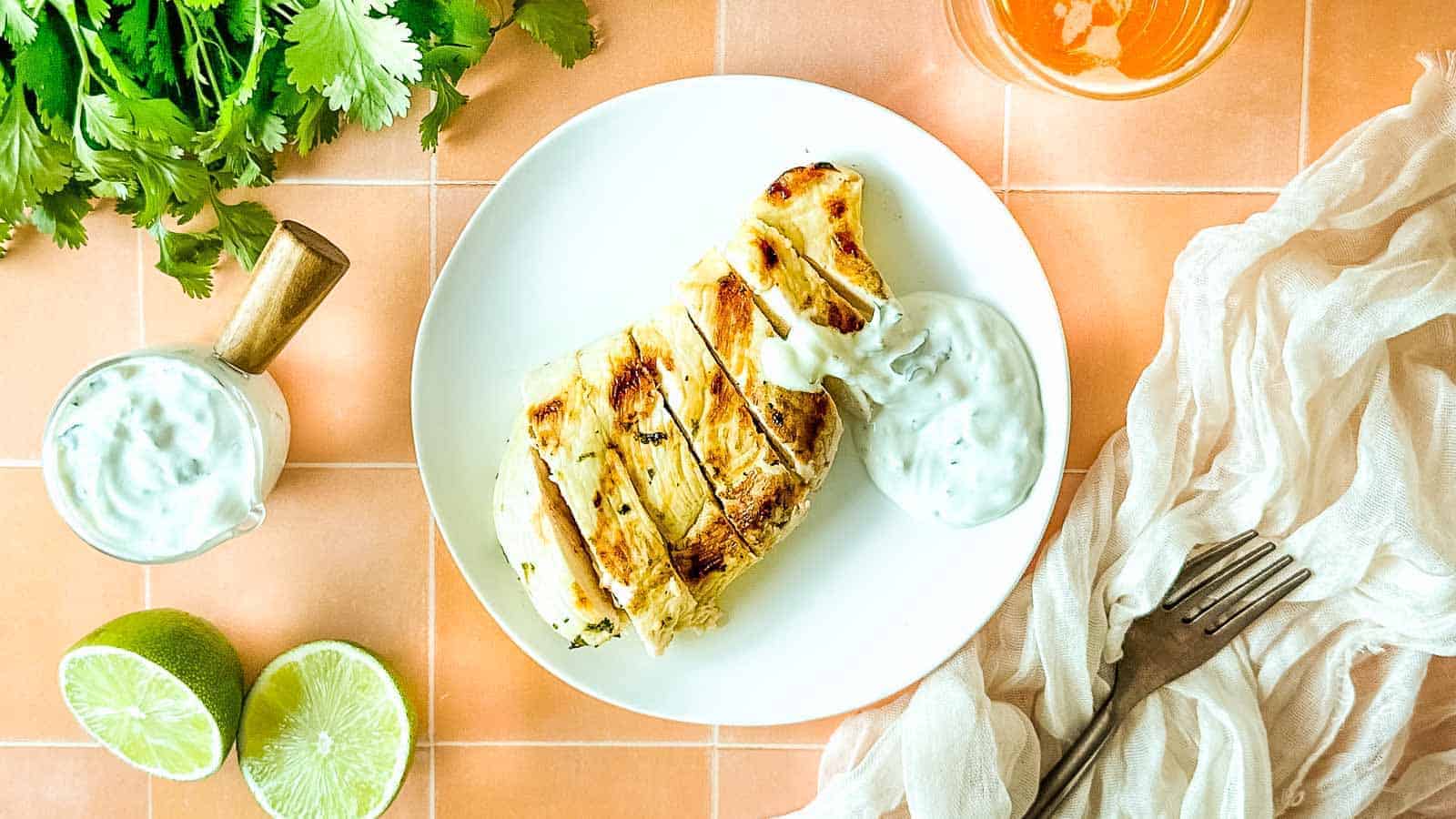 Grilled chicken with cilantro crema on a white plate surrounded by limes, cilantro, and a glass of beer.