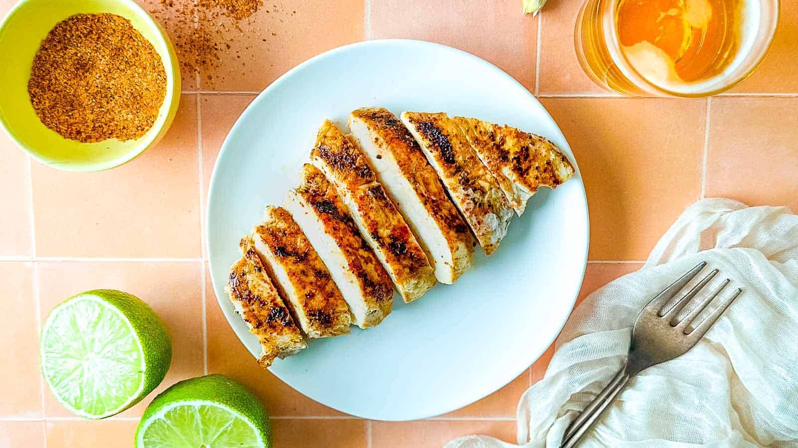 Sliced Tajín Grilled Chicken sits on a white plate surrounded by Tajín seasoning, a sliced lime, a white linen, and a small glass of beer.