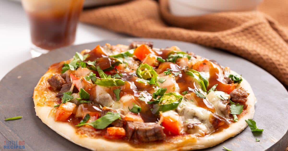 Personal-sized pizza topped with diced tomatoes, leafy greens, and chunks of meat, drizzled with sauce on a dark plate. A drink and a brown cloth are in the background.
