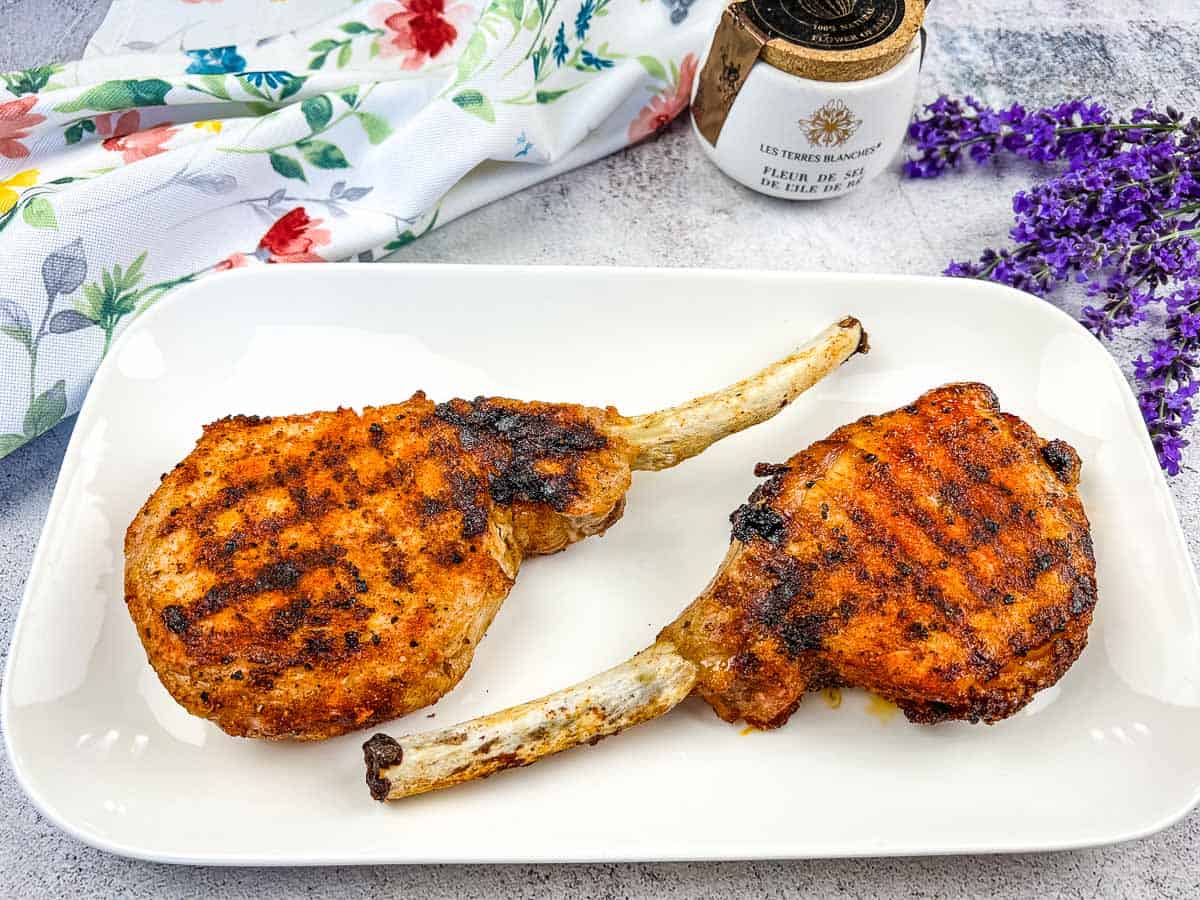 Two grilled seasoned lamb chops on a white plate, garnished with a lavender sprig and accompanied by floral patterned cloth and a spice jar in the background.