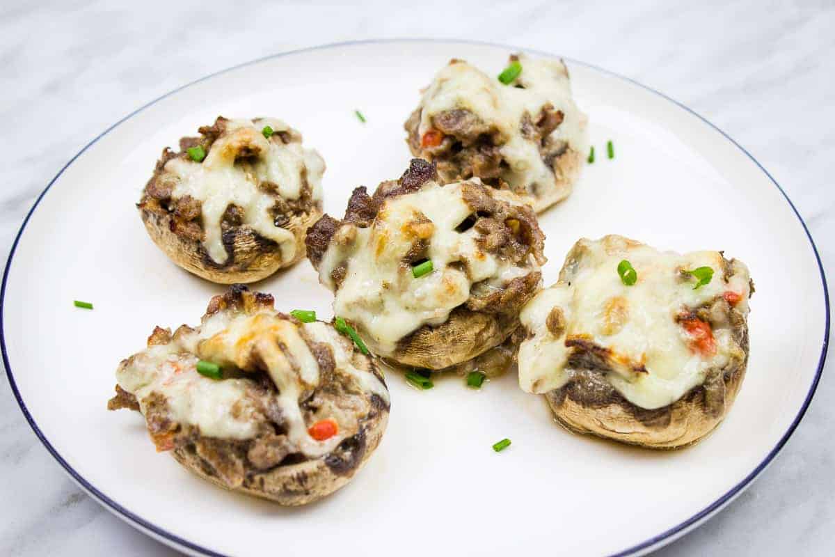 Five stuffed mushrooms topped with melted cheese and garnished with chopped chives on a white plate.