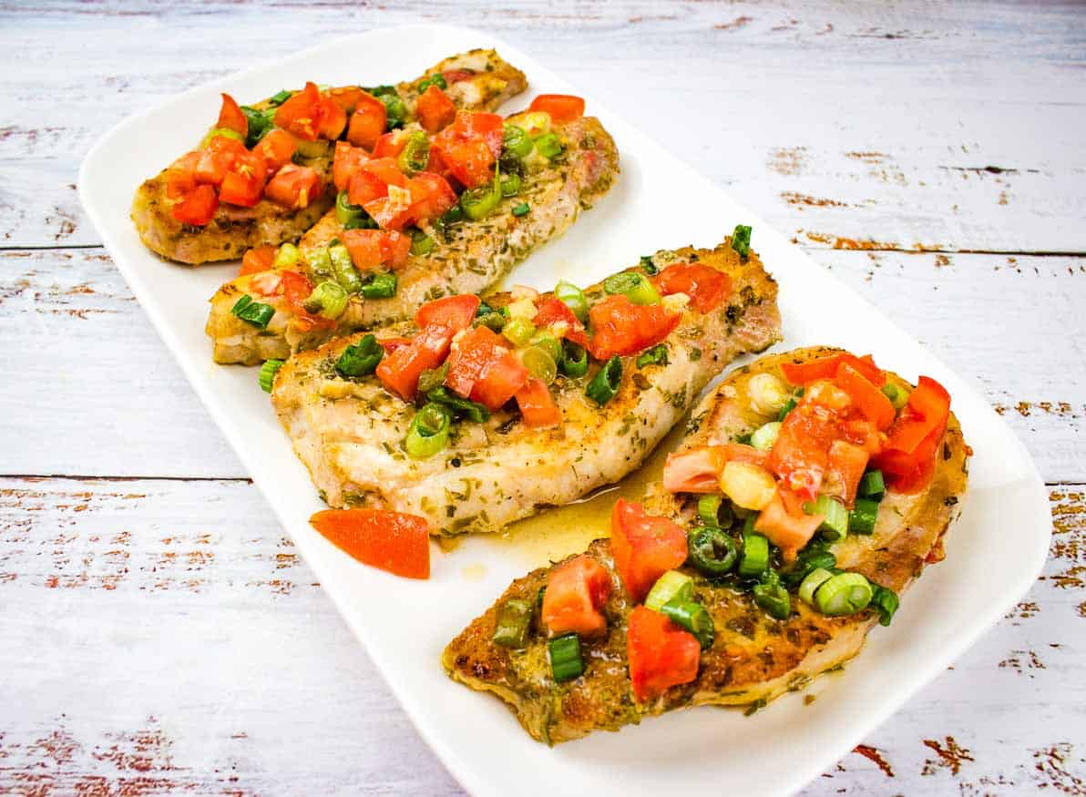 Grilled chicken breasts topped with diced tomatoes and green onions on a rectangular white plate, placed on a light wooden surface.
