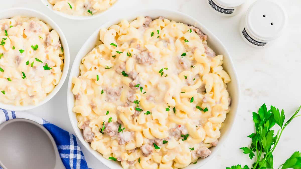 Bowl of creamy macaroni and cheese with ground beef, garnished with chopped parsley. Additional small bowls with the same dish nearby, placed on a white surface with a blue napkin.