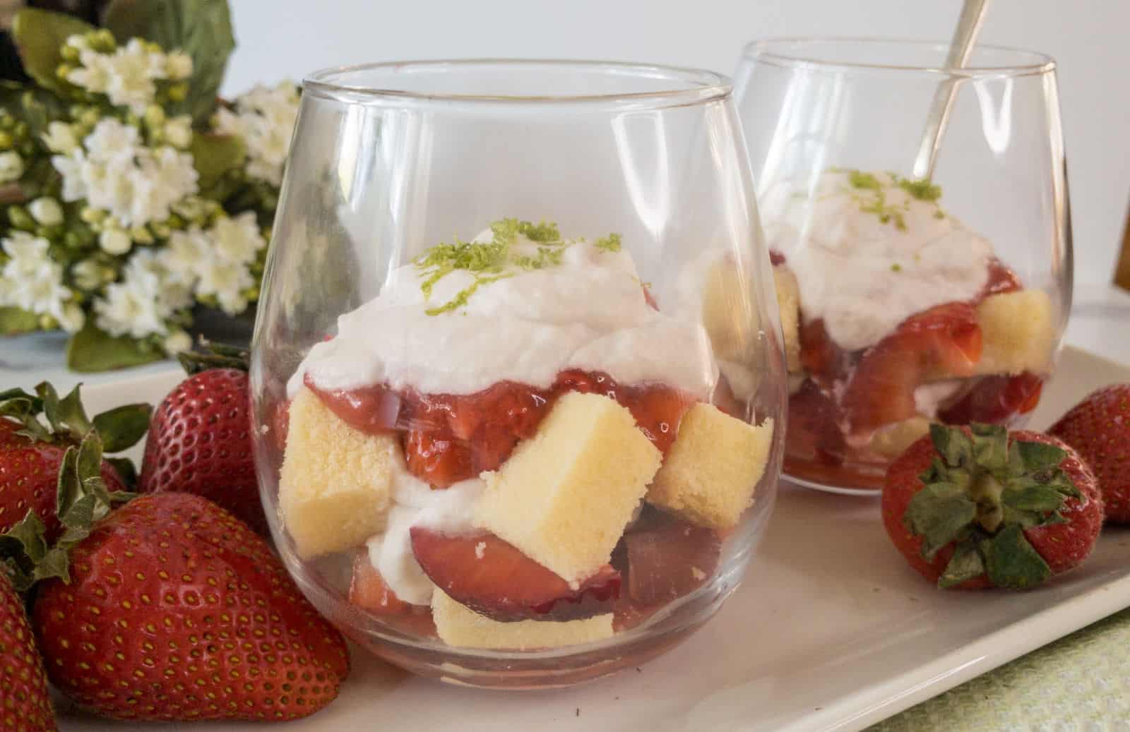 Two glasses filled with layered dessert of sponge cake cubes, strawberries, and whipped cream. A few whole strawberries and a flower arrangement are in the background.