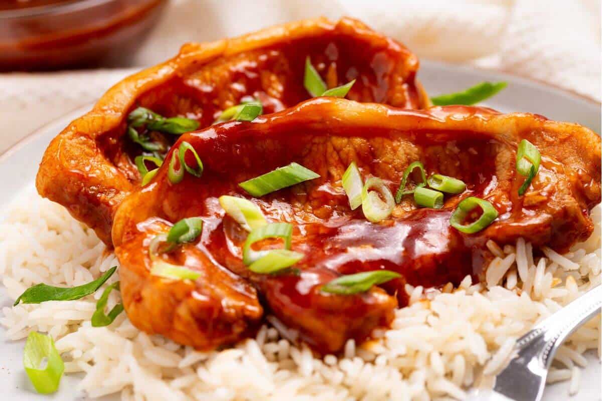 Two pork chops with sauce and green onions on a bed of white rice, served on a white plate.
