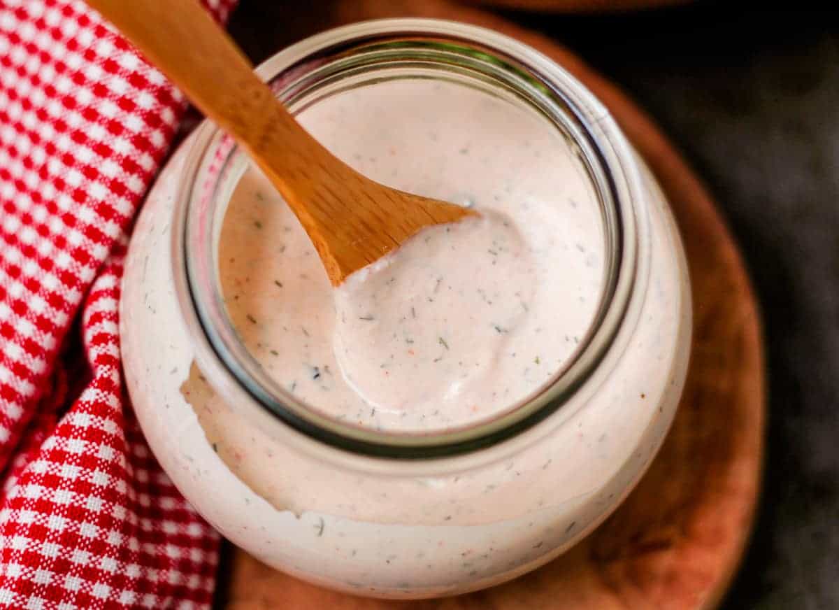 A jar of creamy ranch dressing with visible herbs, accompanied by a wooden spoon and a red checkered cloth.