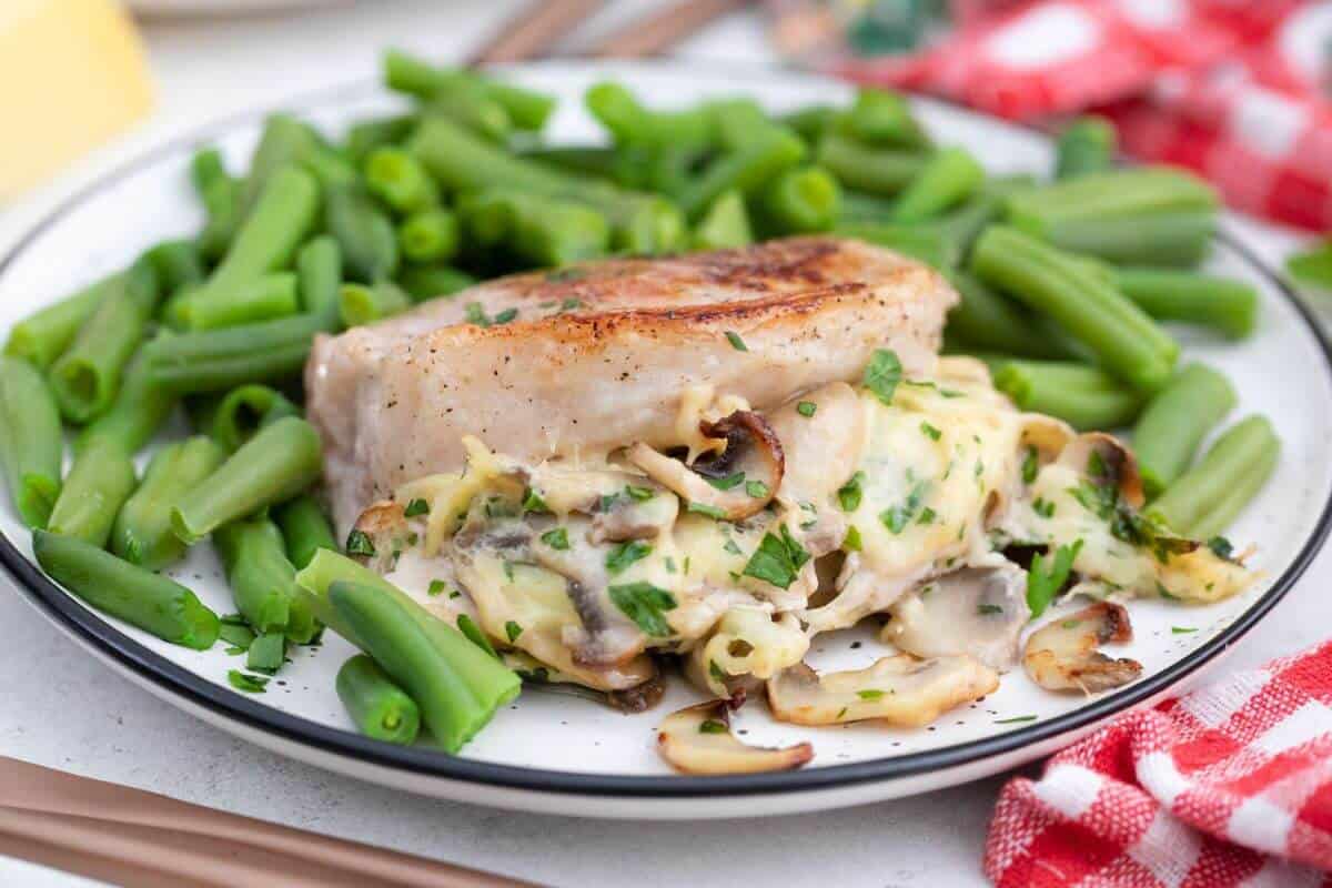 Grilled pork chop topped with mushrooms and herbs, surrounded by cooked green beans on a white plate with a red checkered cloth nearby.