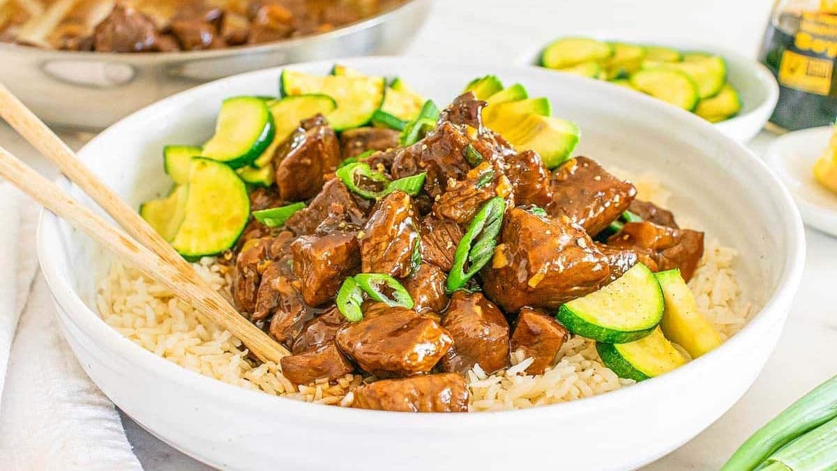 A bowl of beef stir-fry with zucchini and green onions on rice. Chopsticks are placed on the bowls edge.