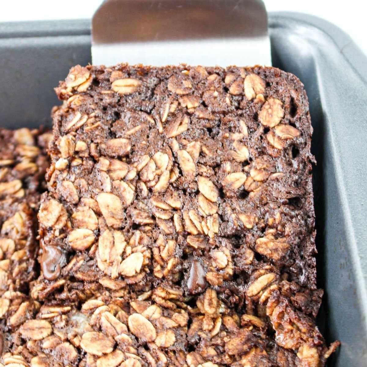 A baked chocolate oatmeal bar being lifted from a baking pan with a metal spatula.