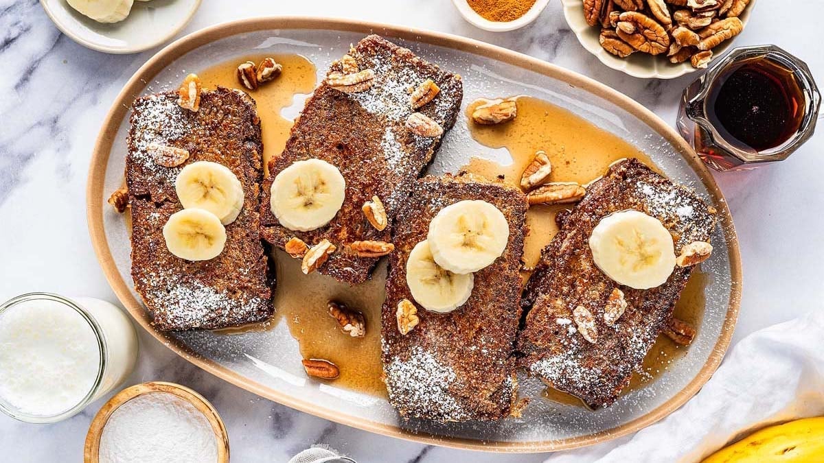 A platter of banana bread french toast with powdered sugar, banana slices, and pecans, drizzled with syrup.