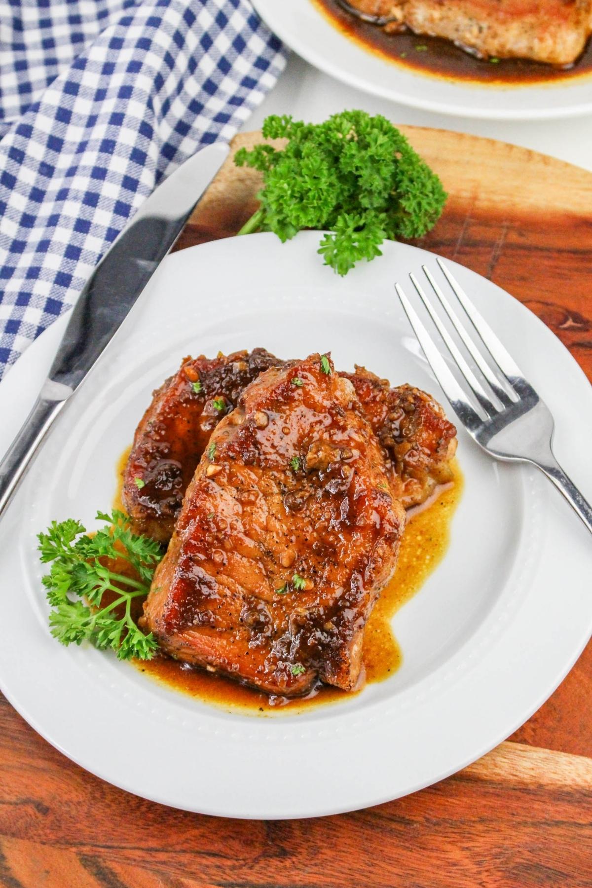 Plate of glazed pork chops garnished with parsley, accompanied by a fork and knife, on a wooden table next to a blue and white checkered cloth.