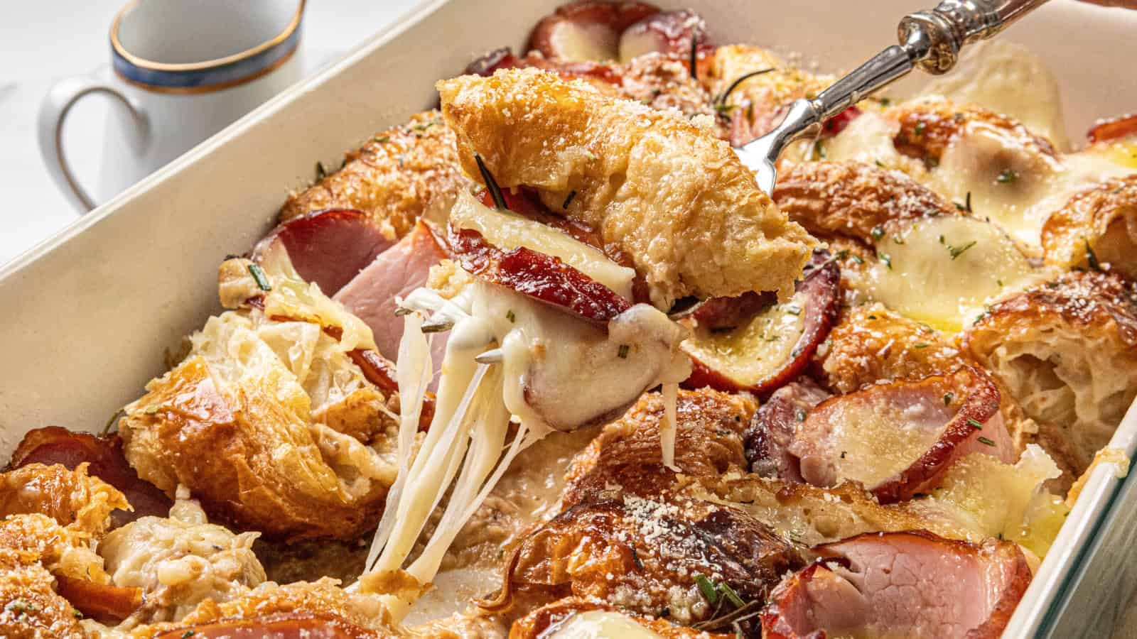 A fork lifts a portion of croissant breakfast casserole with cheese and slices of fruit, from a baking dish. A cup sits in the background.