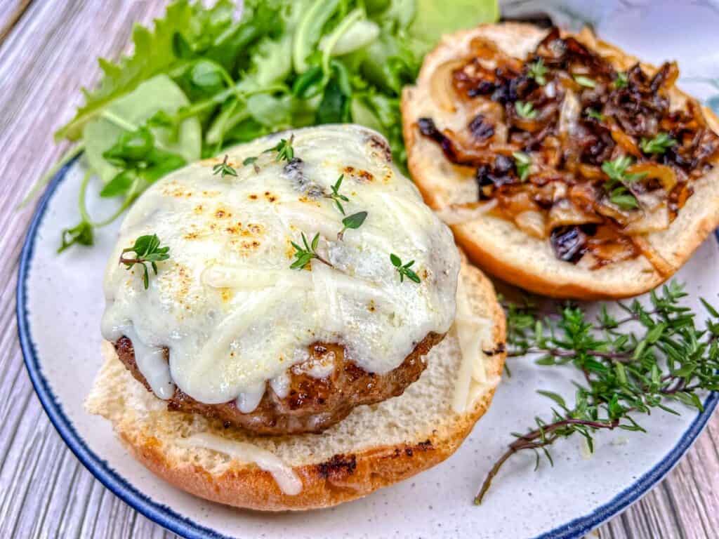 Open sandwich with a beef patty topped with melted cheese and herbs, caramelized onions on the top bun, and mixed greens on the side, served on a plate.