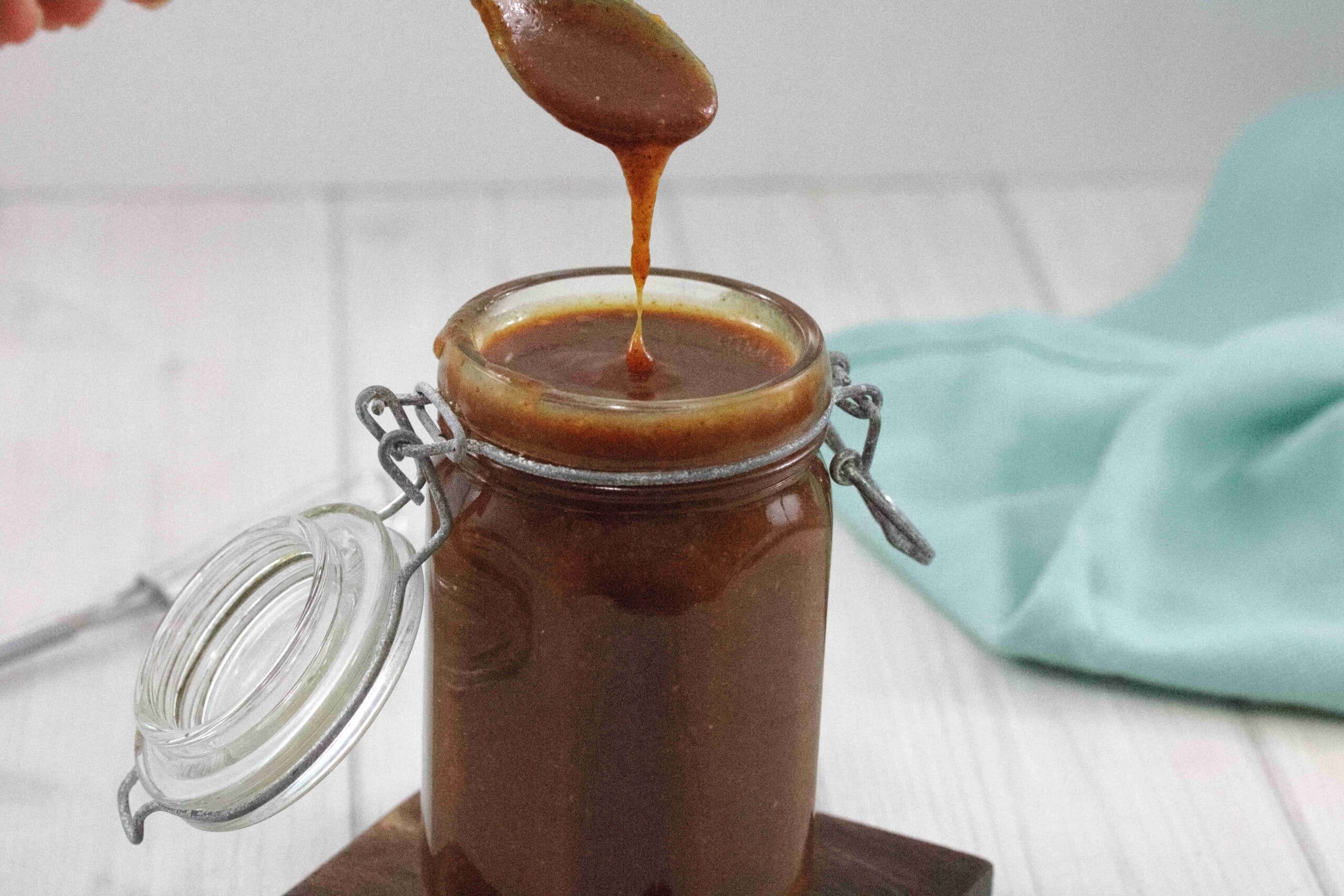 Small mason jar with homemade caramel sauce in it and a spoon pulling out the caramel with a blue linen behind it.